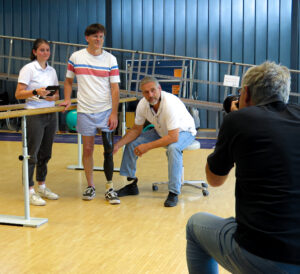 Ein Fotograf macht ein Bild von drei Personen in einem Sanitätshaus. Zwei der Personen tragen Prothesen an den Beinen und stehen an einer Gehstange, während eine Person in weißem Shirt daneben steht. Im Hintergrund sind Turngeräte und eine blaue Wandverkleidung zu sehen. Die Szene findet in einer hellen Halle statt.